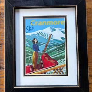 May include: A framed print featuring a vintage-style illustration of a person in a blue jacket and yellow pants riding a red ski lift. The background shows a snow-covered mountain and green trees. The text reads Ski Cranmore in yellow and blue.