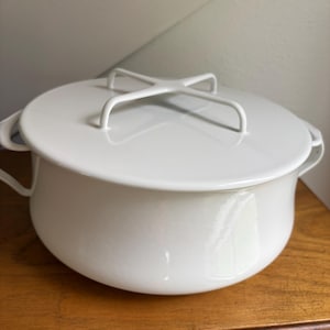 May include: A solid white enameled pot with a matching lid. The lid features a distinctive cross-shaped handle design. The pot has two curved side handles and a rounded body, providing a clean and uniform appearance for this kitchen cookware item.