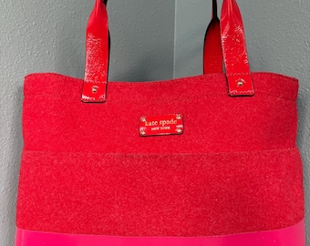 May include: A red tote bag with a felted texture and a glossy pink bottom. The bag has red patent leather handles and a small rectangular label that reads "kate spade new york".