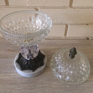 May include: A clear glass candy dish with a diamond pattern and a marble base. The dish has a lid with a small knob on top.