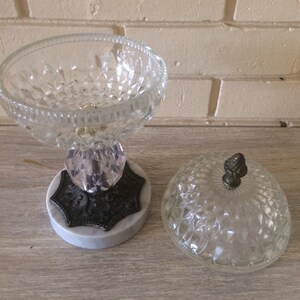 May include: A clear glass candy dish with a diamond pattern and a marble base. The dish has a lid with a small knob on top.