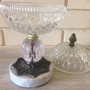 May include: A clear glass candy dish with a marble base and a matching lid. The dish has a decorative, faceted design and a metal base with an inscription.