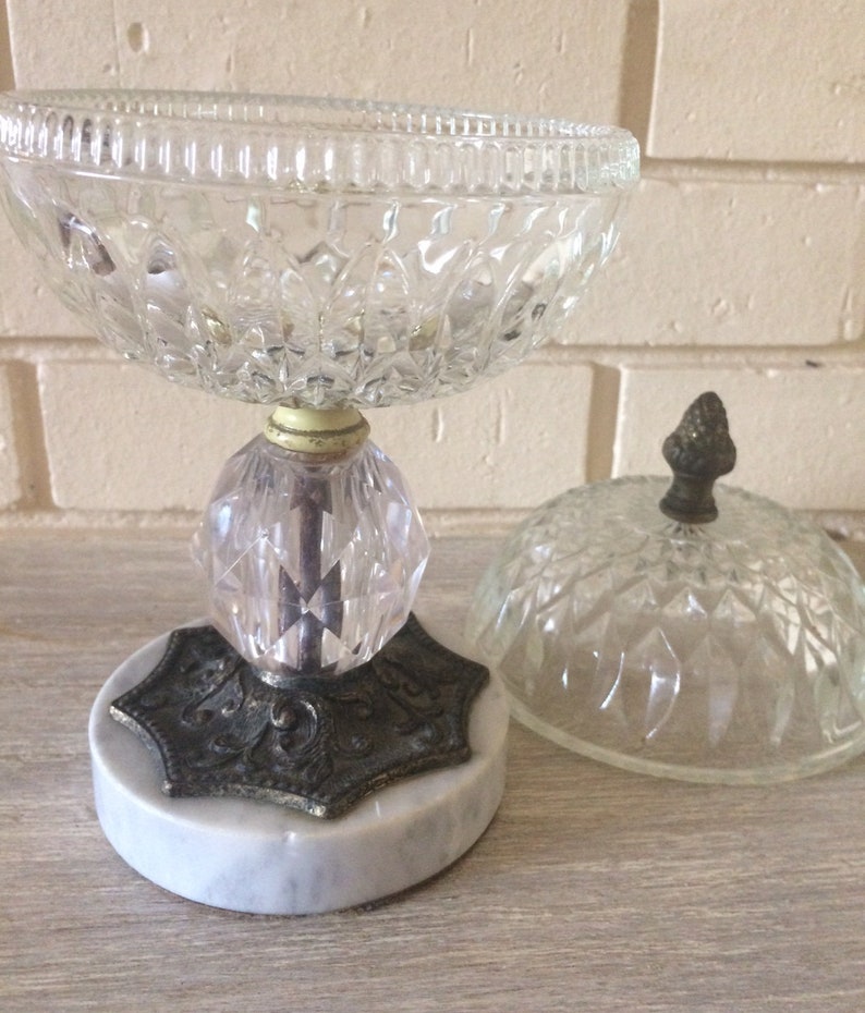 May include: A clear glass candy dish with a marble base and a matching lid. The dish has a decorative, faceted design and a metal base with an inscription.