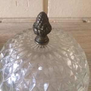 May include: A clear glass dome with a decorative, ornate, metal finial. The dome has a diamond-shaped pattern.