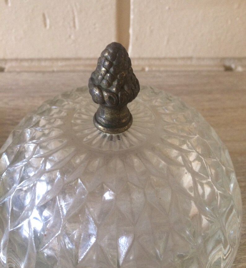 May include: A clear glass dome with a decorative, ornate, metal finial. The dome has a diamond-shaped pattern.