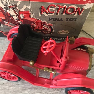 May include: Red toy car with a black seat and a steering wheel. The car is labeled "ACTION PULL TOY" on the box.
