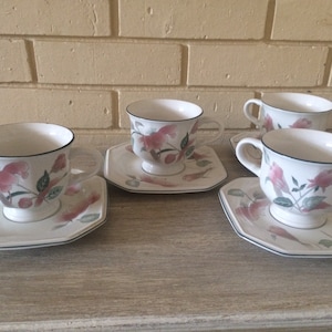 May include: Set of four white teacups and saucers with a floral design featuring pink and green flowers. The cups and saucers have a black rim.