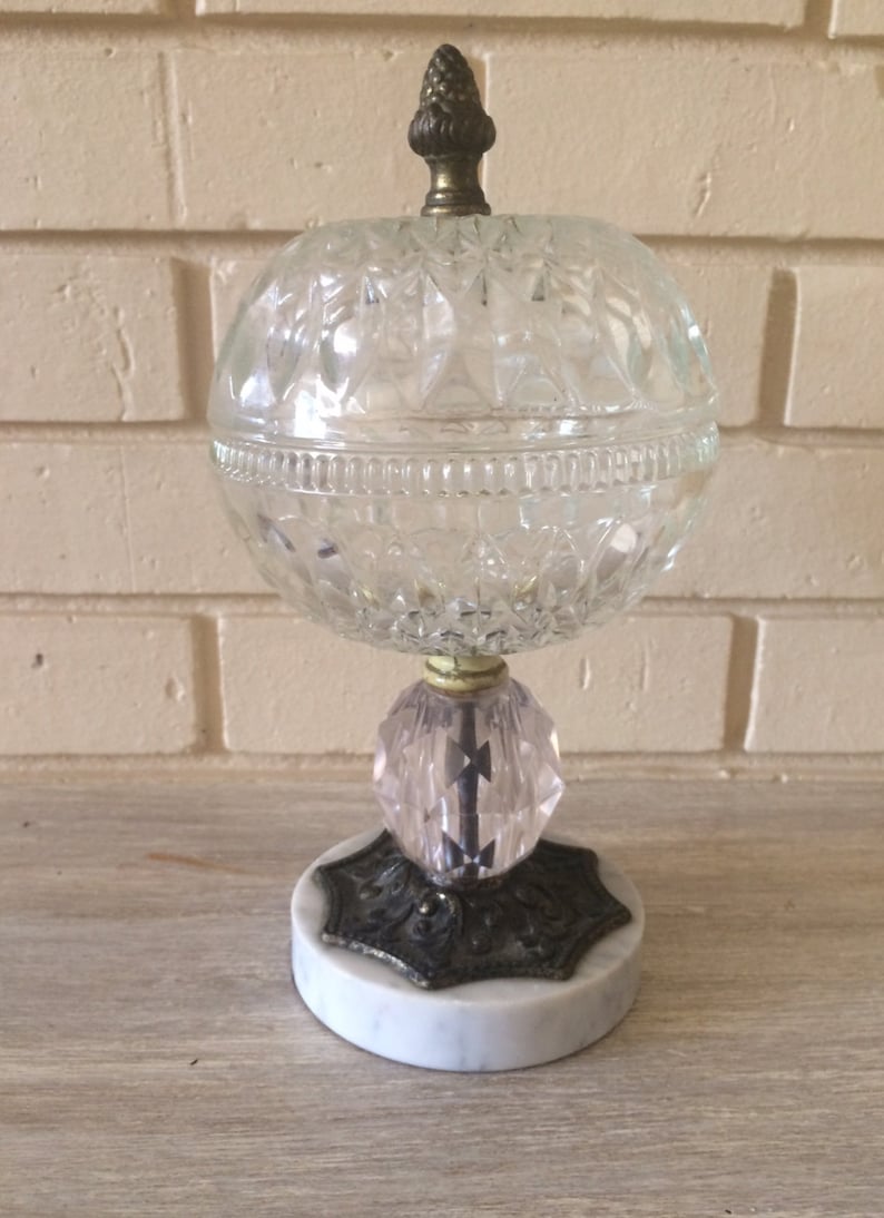 May include: A clear glass globe with a cut glass design sits on a pink glass base with a metal and marble base. The globe has a small metal finial on top.
