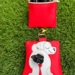 May include: Red dog waste bag dispenser with a white dog design. The dispenser has a zip closure and a gold-coloured clip. Includes a roll of black waste bags.