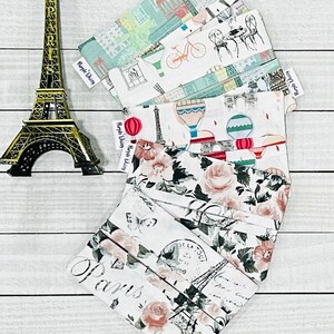 May include: A stack of five fabric pouches featuring various Parisian-themed prints. The pouches are white with different designs including the Eiffel Tower, hot air balloons, bicycles, and floral patterns. Each pouch has a small purple label that says "Purple Daisy".