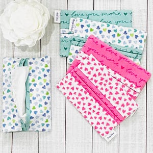 May include: A set of three tissue box covers with a heart pattern. The covers are made of fabric and have a light blue, green, and pink colour scheme. The covers feature the text "love you more" and "love you".