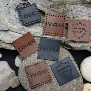 Può includere: Sette etichette in pelle con diversi testi. Le etichette sono marroni e nere e hanno un bordo cucito. Le etichette sono disposte su un pezzo di legno di deriva e alcuni sassi.