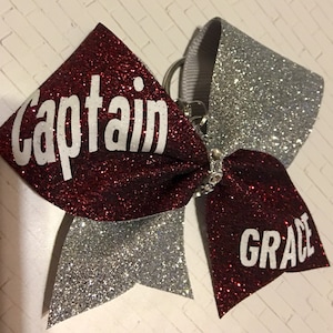 May include: A red and silver glitter cheer bow with a white keychain and the words "Captain" and "GRACE" printed on it.