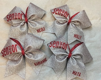May include: Four silver glitter cheer bows with red accents. Each bow has the word "Senior 2018" and a name: Nicole, Molly, Karianna, and Maya.