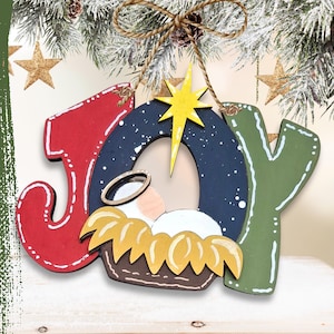 May include: A wooden Christmas ornament spelling "JOY" with a nativity scene. The letters are red and green with white accents. The nativity scene features a baby in a manger, a star, and a blue night sky.