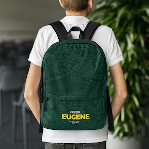 May include: Green backpack with a detailed black line drawing of a stadium and surrounding trees. The text "OREGON EUGENE" is in yellow, with a small signature below. The backpack has black straps and a black top handle.