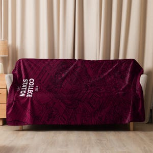 May include: A maroon fleece throw blanket featuring a black and white outline of a city map. The text "College Station" is printed in white on the blanket.