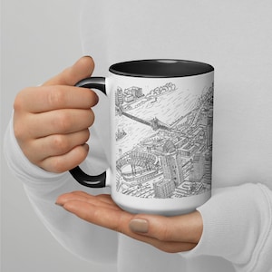 May include: A white ceramic mug with a black rim and handle. The mug features a black and white line drawing of a city skyline with a bridge and a stadium.