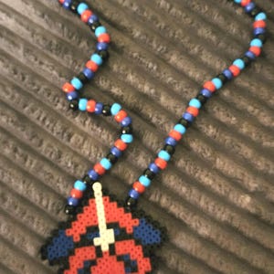 May include: A beaded necklace featuring a pendant of a superhero's face. The pendant is red, black, blue, and white. The necklace chain is made of red, blue, and black beads. The necklace is on a gray surface.
