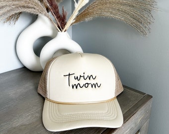 May include: A beige trucker hat with the text "Twin mom" in black script. The hat has a mesh back and a curved brim. A gold-colored rope detail accents the front. The hat is displayed on a wooden surface with a white vase and dried flowers in the background.