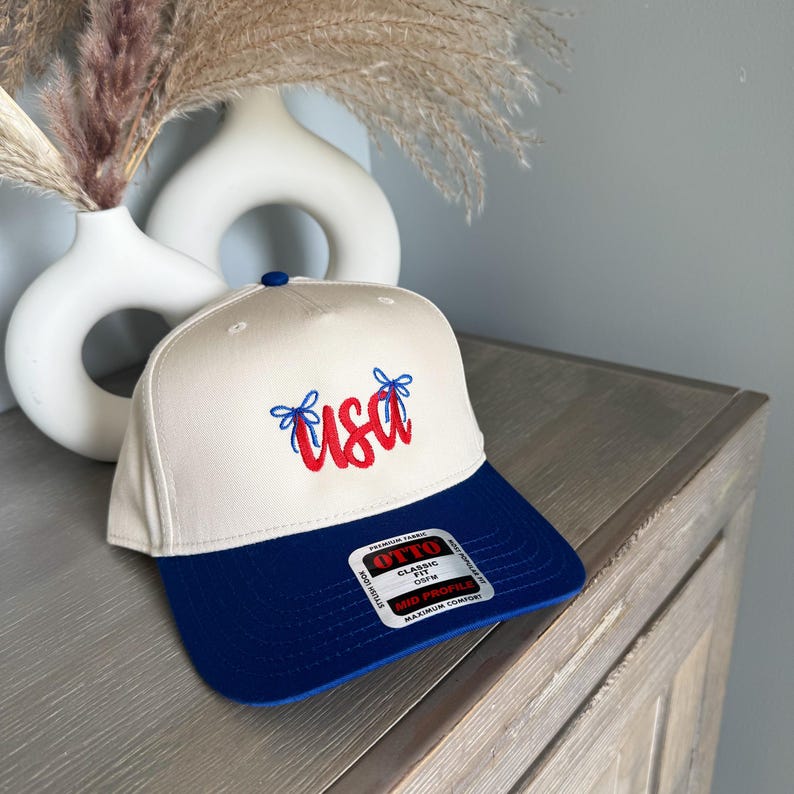 May include: A cream and navy blue baseball cap with the word "USA" embroidered in red script. The cap has a navy blue brim and a blue button on top. The hat is on a wooden surface.