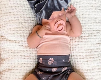May include: A baby wearing a gray velvet turban, a pink tank top, black and gray patterned bloomers, and gray leg warmers. The baby is wearing a pink pacifier.