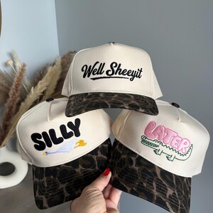May include: Three cream-colored baseball caps with leopard print brims. The top cap features black embroidered text reading Well Sheeyit. The bottom left cap has black embroidered text SILLY with a goose. The bottom right cap has pink text LATER with a green alligator.