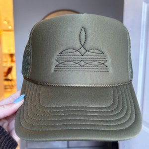 Puede incluir: Una gorra de camionero verde con un dise&ntilde;o geom&eacute;trico bordado en negro en la parte delantera. La gorra tiene una parte trasera de malla y una visera curvada.