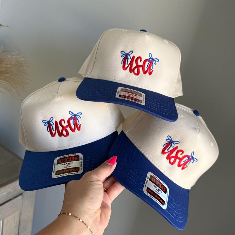 May include: Three baseball caps in cream and blue, each embroidered with the word "USA" in red. The caps have a blue peak and are held together. The caps are a classic style.