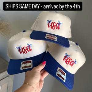 May include: Three baseball caps in cream and blue, each embroidered with "USA" in red script. The caps have a blue peak and a side patch. The text "SHIPS SAME DAY - arrives by the 4th" is at the top.