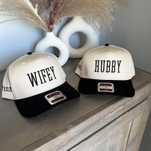 May include: Two baseball caps with black brims and cream-colored crowns. One cap says "WIFFY" in black letters, the other says "HUBBY". Both caps have a black button on top and a tag on the brim.