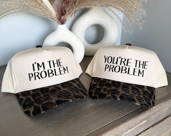 May include: Two white trucker hats with leopard print visors. One hat reads "I'M THE PROBLEM" and the other reads "YOU'RE THE PROBLEM" in black text. The hats are displayed on a wooden surface.
