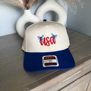 May include: A beige and blue baseball cap with the word "USA" embroidered in red script. The cap has a blue brim and a blue button on top. The text "OTTO" is visible on a tag.