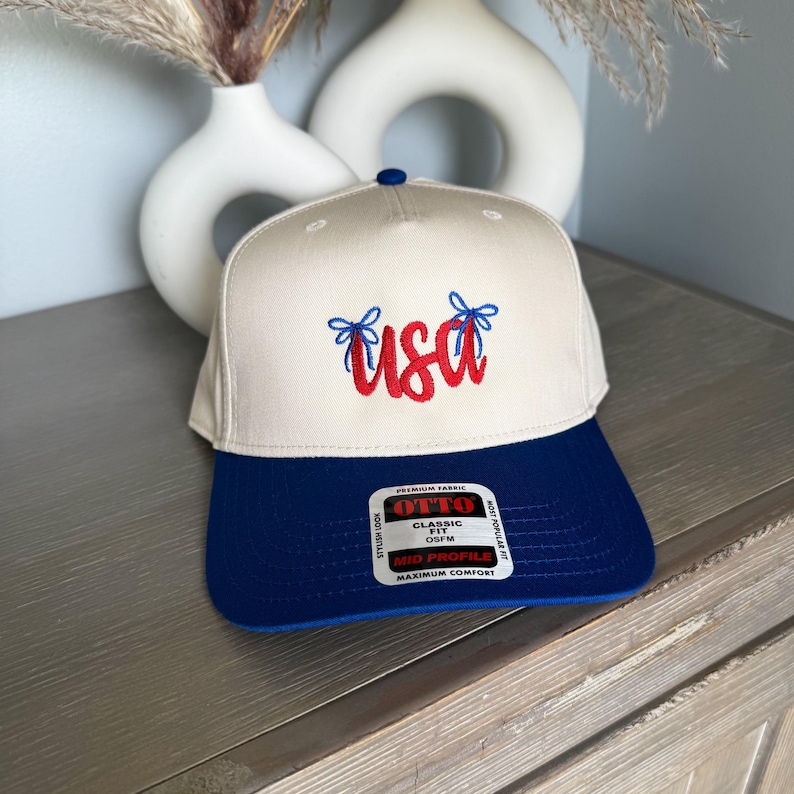 May include: A beige and blue baseball cap with the word "USA" embroidered in red script. The cap has a blue brim and a blue button on top. The text "OTTO" is visible on a tag.