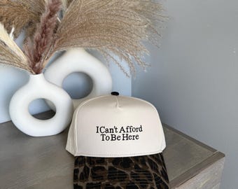 May include: A cream-colored baseball cap with the text "I Can't Afford To Be Here" in black. The brim features a brown and black leopard print. The cap is set on a wooden surface with a white vase and decorative dried plants in the background.