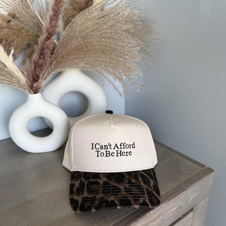 Puede incluir: Gorra de b&eacute;isbol color crema con el texto "I Can't Afford To Be Here" en negro. La visera presenta un estampado de leopardo marr&oacute;n y negro. La gorra est&aacute; colocada sobre una superficie de madera con un jarr&oacute;n blanco y plantas secas decorativas en el fondo.