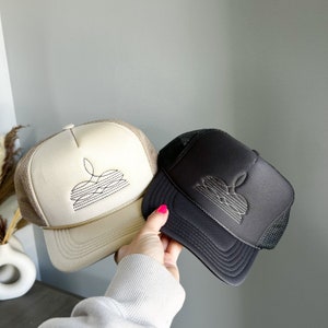Puede incluir: Dos gorras de camionero, una beige y otra negra, con un dise&ntilde;o gr&aacute;fico de una flor estilizada en cada una. La gorra beige tiene un dise&ntilde;o negro y la gorra negra tiene un dise&ntilde;o blanco.