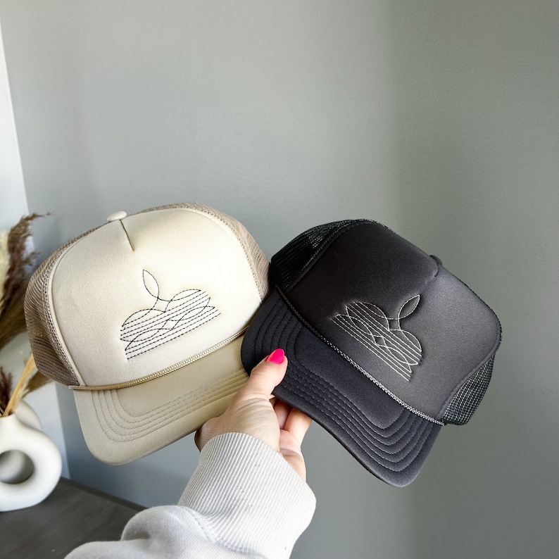Puede incluir: Dos gorras de camionero, una beige y otra negra, con un dise&ntilde;o gr&aacute;fico de una flor estilizada en cada una. La gorra beige tiene un dise&ntilde;o negro y la gorra negra tiene un dise&ntilde;o blanco.