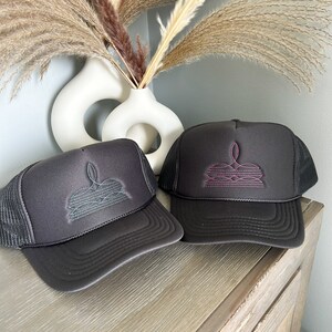 Puede incluir: Dos gorras de camionero negras con paneles traseros de malla. Una gorra tiene un dise&ntilde;o bordado en turquesa y la otra tiene un dise&ntilde;o bordado en rosa.