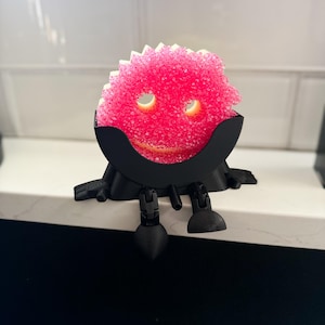 May include: A whimsical, pink, scrubby sponge with a smiling face, held in a black, decorative holder with legs. The sponge has two eye holes and a textured surface, creating a playful, cartoon-like appearance. The holder and legs are made of black plastic.