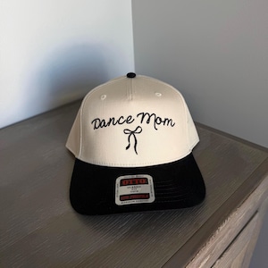 May include: A baseball cap with a cream-colored crown and a black brim. The words "Dance Mom" are embroidered in black script on the front, with a small bow detail. The cap is made of a premium fabric and has a classic fit.