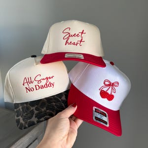 May include: Three baseball caps with different designs. One is white with a red brim and the words "Sweet Heart" in red script. Another is white with a leopard print brim and the words "All Sugar No Daddy" in red script. The third is white with a red brim and a cherry design.