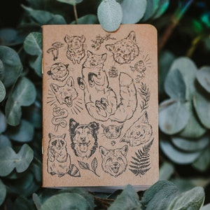 Puede incluir: Un cuaderno marrón con una variedad de ilustraciones de animales, incluyendo zorros, mapaches y una serpiente. Las ilustraciones son en blanco y negro y están rodeadas de hojas y otros diseños inspirados en la naturaleza.