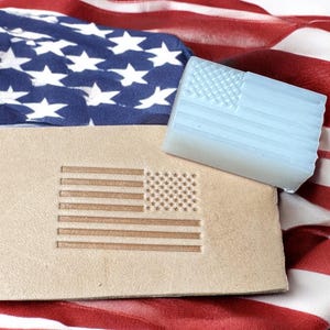 Puede incluir: Un sello de cuero con un diseño de bandera estadounidense marrón sobre un fondo beige. El sello está sobre una bandera estadounidense roja, blanca y azul.