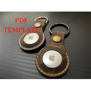 May include: Two brown leather keychains with a silver Apple logo on each. The keychains are stitched with white thread and have a gold metal ring.