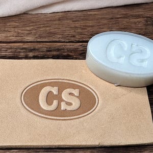 Peut inclure: Un tampon ovale blanc avec les lettres "CS" en relief. Le tampon est posé sur un morceau de cuir brun clair avec les mêmes lettres en relief.