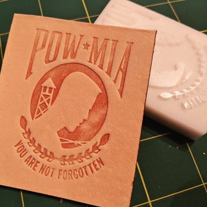 May include: A light brown leather square with the words "POW*MIA" and "YOU ARE NOT FORGOTTEN" embossed in a darker shade. The design includes a silhouette, a watchtower, and a wreath. A white mold is next to the leather.