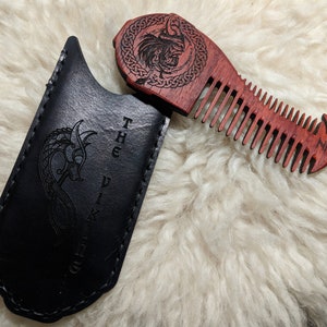 May include: A wooden comb with a Viking design, featuring a dragon and a knotwork pattern. The comb is red and brown and has a black leather case with the words "The Vikings" engraved on it.