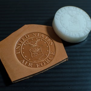 May include: A brown leather patch with a United States Air Force seal embossed on it. The seal features a winged eagle, a star, and the text "UNITED STATES AIR FORCE" around the perimeter.  The seal is surrounded by a circle of stars.  The text "MCMXLVII" is visible below the eagle.  A white rubber stamp with the same seal is shown next to the leather patch.