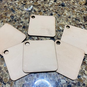 Puede incluir: Seis etiquetas cuadradas de madera sin terminar con un agujero en la esquina superior. Las etiquetas son de color marrón claro y tienen una superficie lisa.